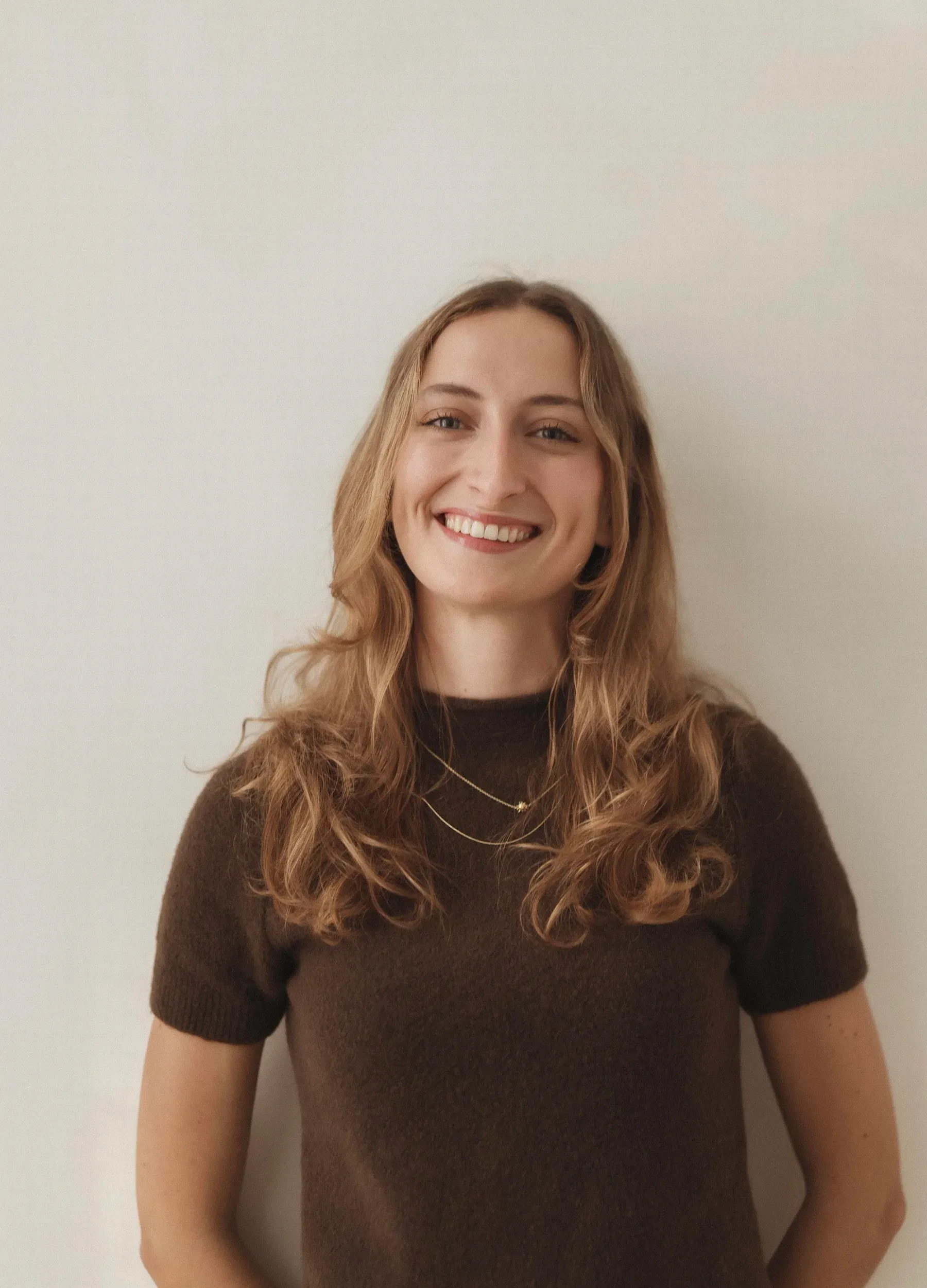 Portrait of Zeliha Stefanie Roesler, smiling, in a brown knit top against a light wall.